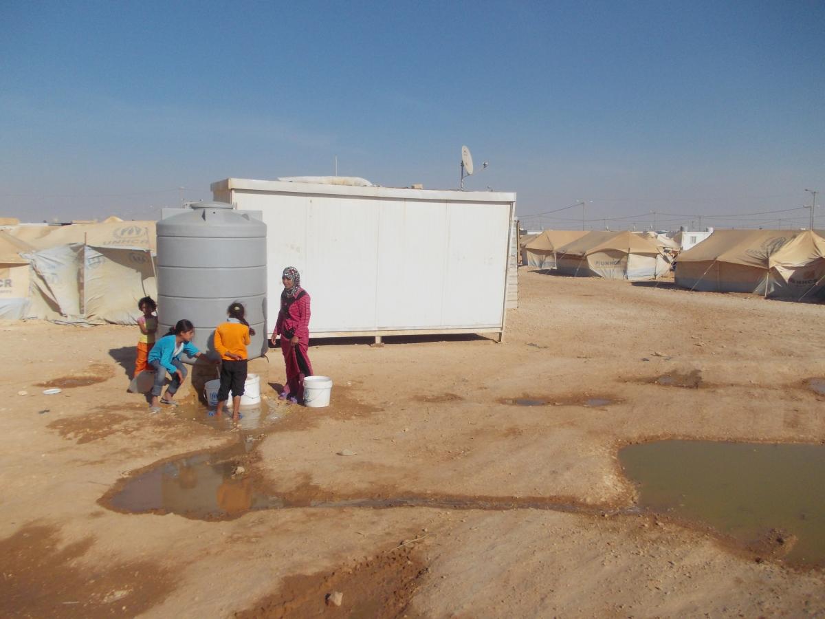 A woman and young girls near a water tank in a Syrian refugee camp